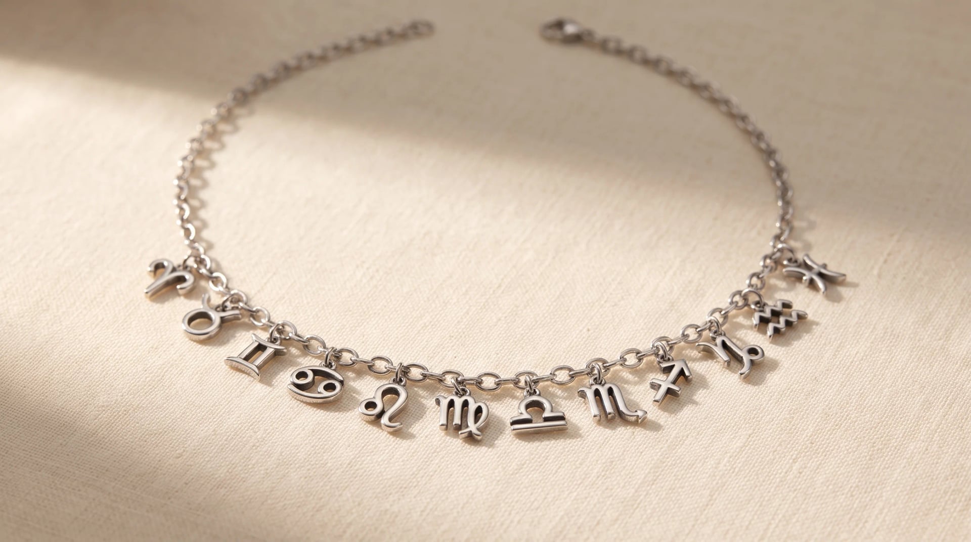 A silver charm necklace with all twelve zodiac symbols arranged on a cream linen surface in soft light, by Enleira